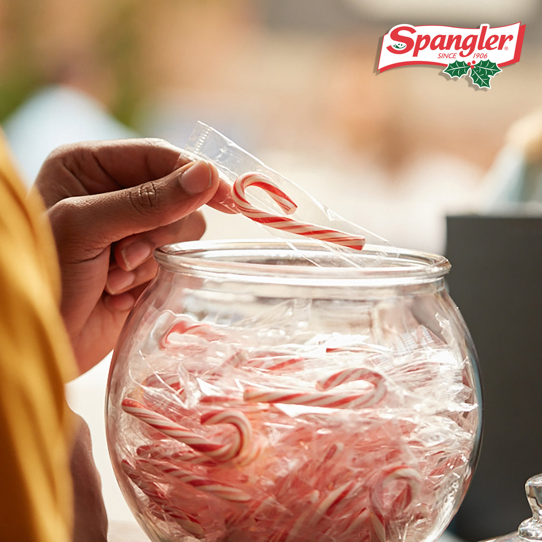 A left hand selecting an individually wrapped Spangler mini candy cane from a clear bowl.